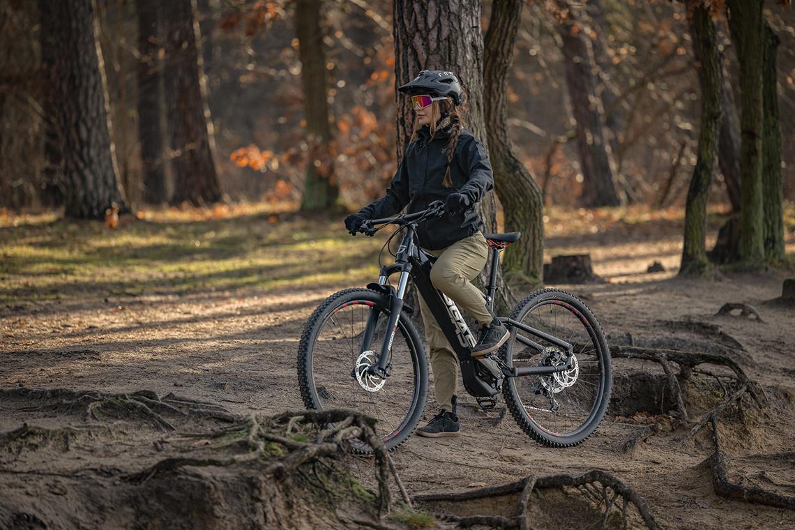 Rowerzystka stoi z rowerem elektrycznym na zboczu pełnym korzeni.